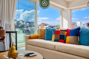Une pièce comprenant un canapé, une table avec un vase et des télécommandes, et offrant une vue sur le paysage urbain à travers de grandes fenêtres.