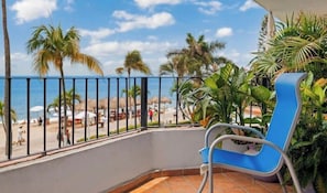 A balcony with a blue chair, a view of the beach, and palm trees.