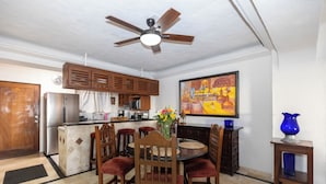 A dining area with a round table, wooden chairs, a ceiling fan, a kitchen in the background, and a painting on the wall. - Ocean View 2Br Condo-balcony-beach-pool - 212 (Puerto Vallarta)
