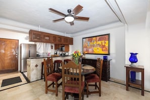 A dining area with a round table, wooden chairs, a ceiling fan, a kitchen in the background, and a painting on the wall.