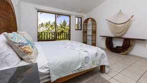 A bedroom with a bed, a hammock, a wooden shelf, and a view of palm trees.