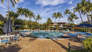 A pool area with lounge chairs and umbrellas, surrounded by palm trees and a clear sky.