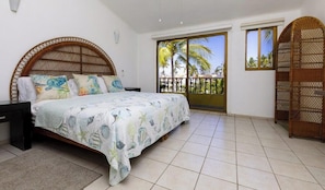 A bedroom with a bed, a nightstand, a window with a view of palm trees, and a balcony with a wicker chair.