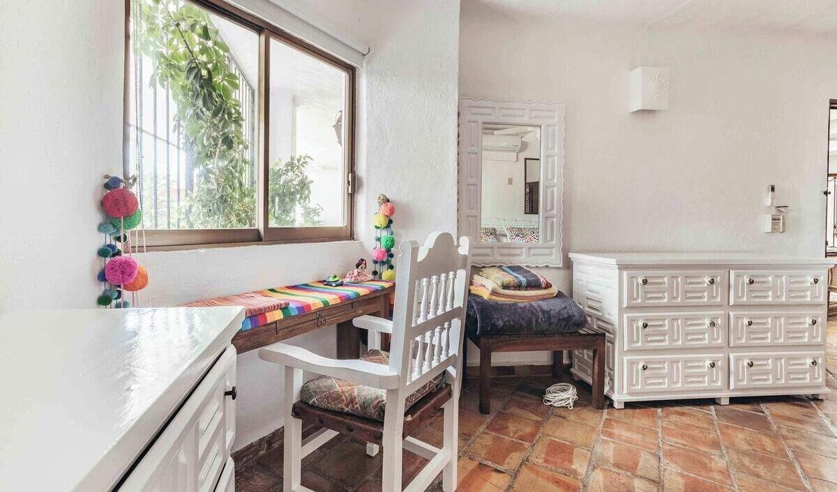 A room with a bed, a desk, a dresser, a window with a view of greenery, and a mirror.