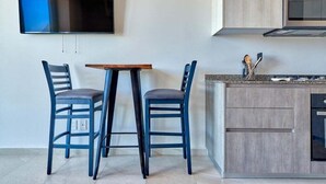 A modern kitchen with a flat-screen TV mounted on the wall, a wooden table, two blue chairs, and a built-in oven.