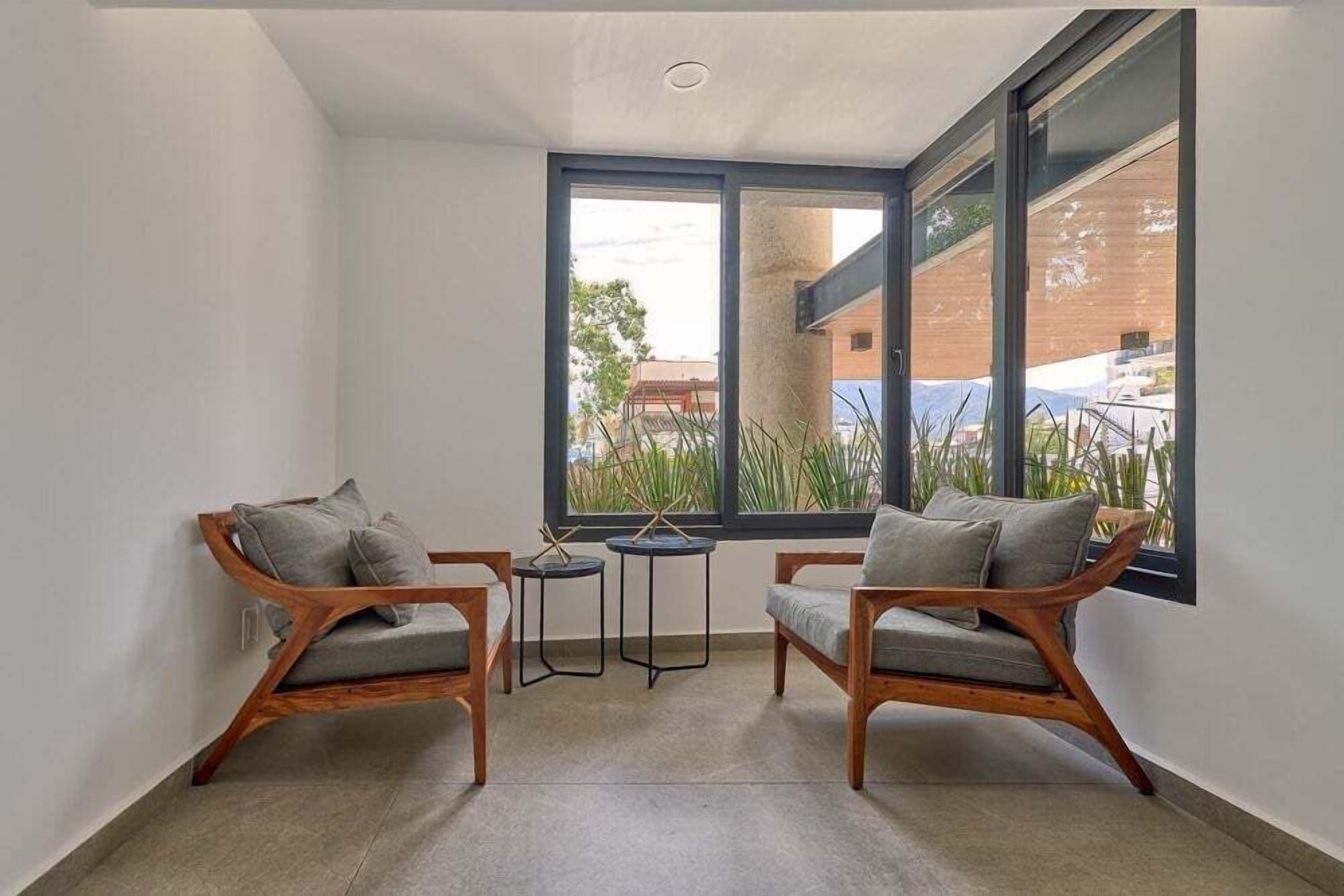 A room with two wooden chairs, a small round table, and a large window with a view of buildings and greenery.