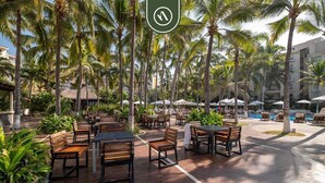 An outdoor dining area with wooden tables and benches, surrounded by palm trees and a pool. - 3BR Condo in the Krystal Grand Hotel - Beach (Nuevo Nayarit)
