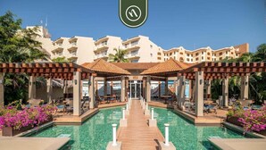 A resort pool area with a wooden walkway leading to a covered seating area, surrounded by buildings with balconies. - 3BR Condo in the Krystal Grand Hotel - Beach (Nuevo Nayarit)
