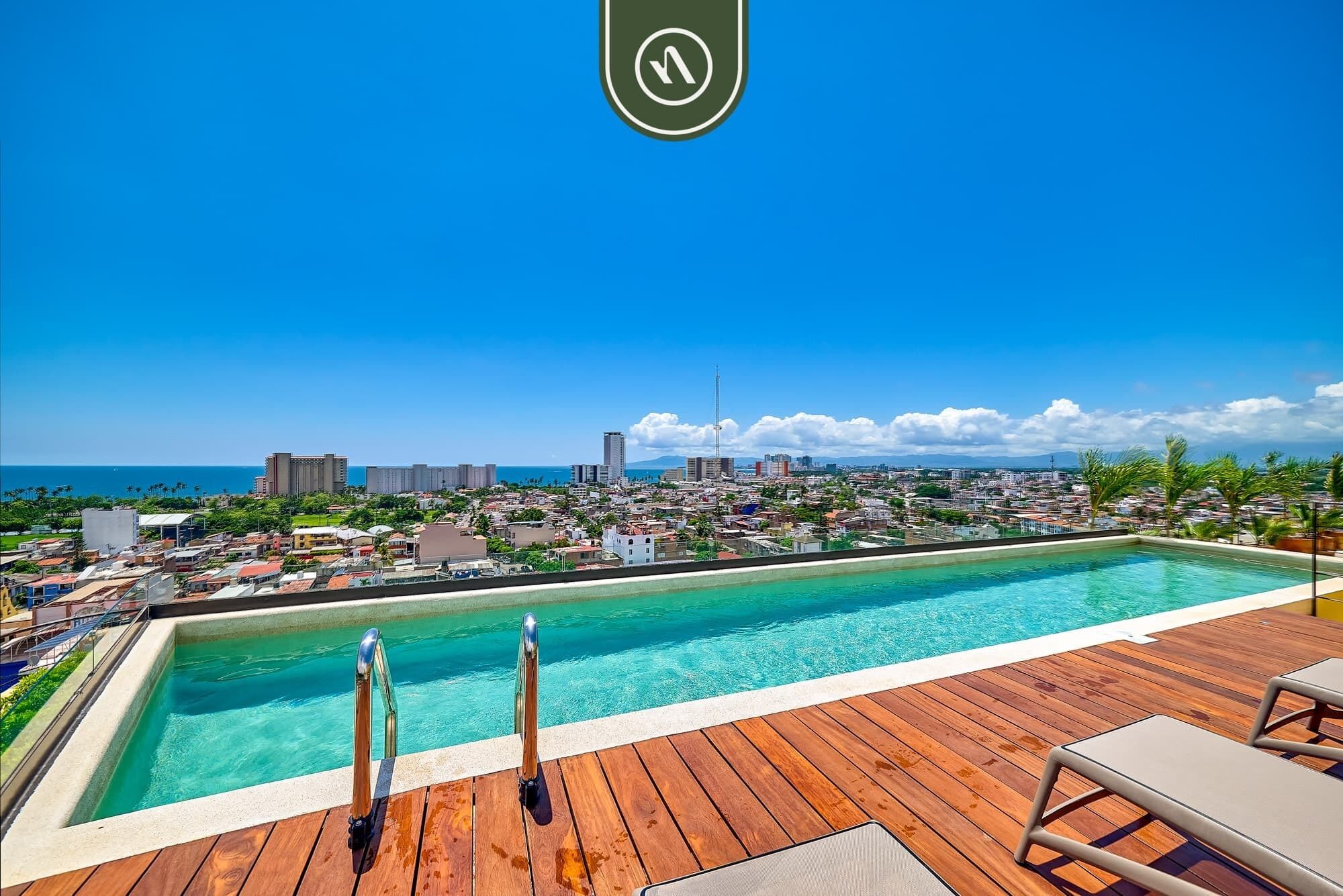 Une piscine dotée d’une terrasse en bois et de chaises longues, offrant une vue sur le paysage urbain et l’océan.