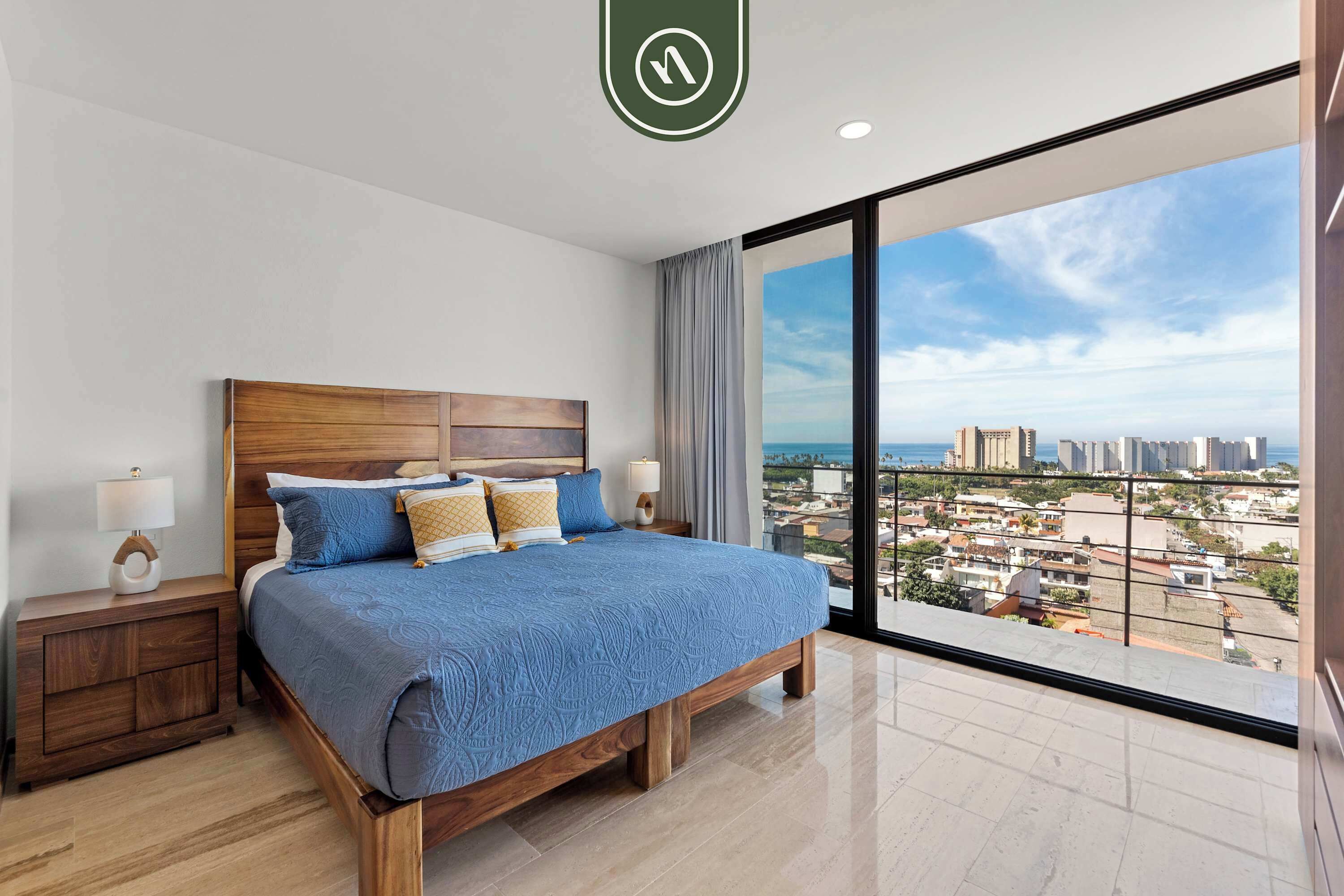 A spacious bedroom with a large bed, wooden headboard, and a city view through floor-to-ceiling windows.