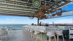 Un bar avec vue sur l’océan, des places assises en plein air et un barman.