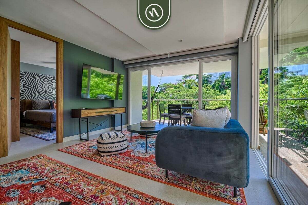 Ein modernes Wohnzimmer mit einem blauen Sofa, einem an der Wand befestigten Fernseher und einem großen Fenster mit Blick auf Grünflächen.