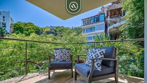 Ein Balkon mit zwei Stühlen und einem Tisch, umgeben von einem Metallgeländer, mit Blick auf eine üppige grüne Landschaft und Gebäude.