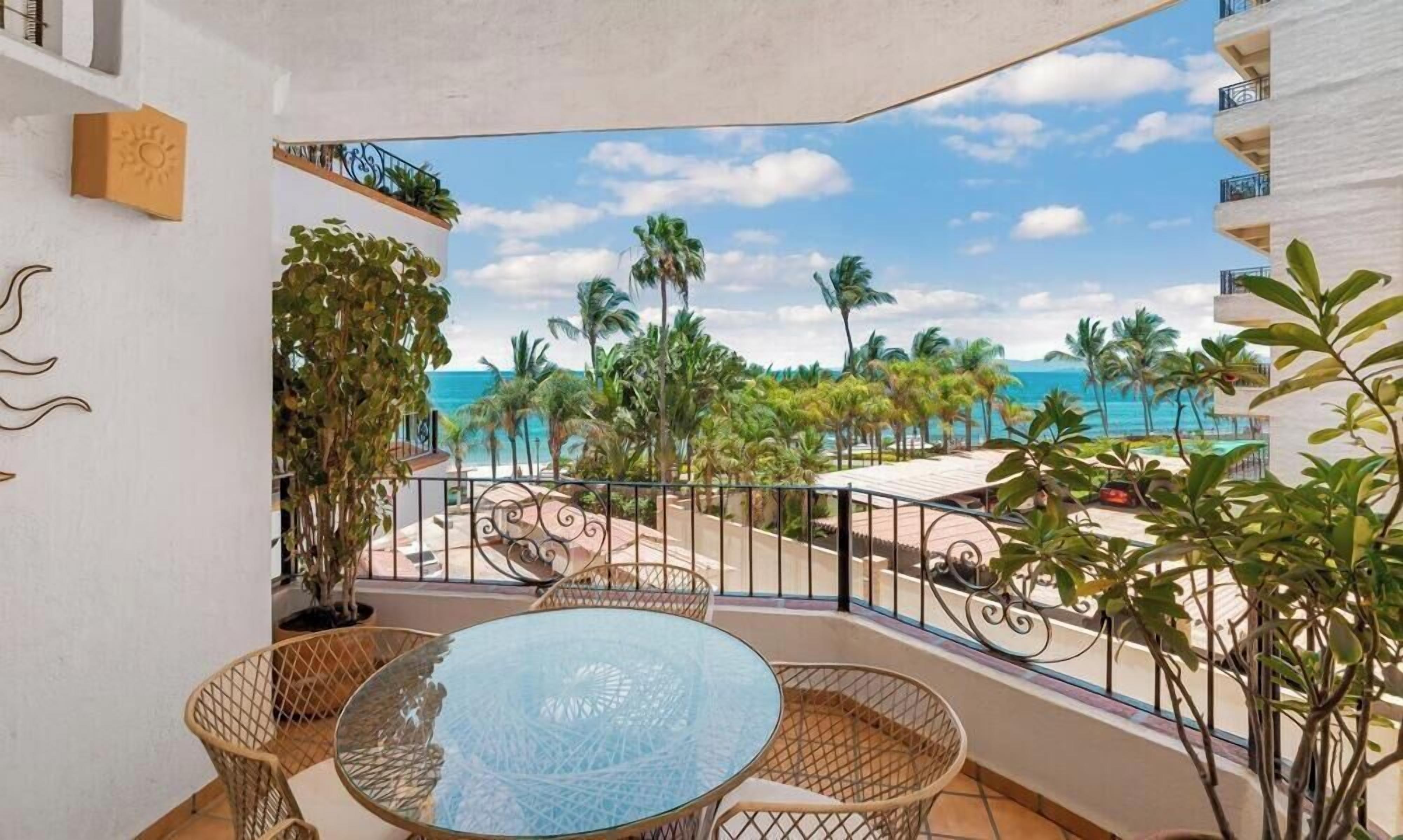 A balcony with a glass table and wicker chairs, overlooking a beach with palm trees and a clear sky.