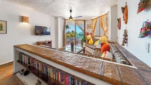 A living room with a long wooden bookshelf, a television, a ceiling fan, and a view of palm trees. - Ocean View 2Br Condo-balcony-beach-pool - 304 (Puerto Vallarta)