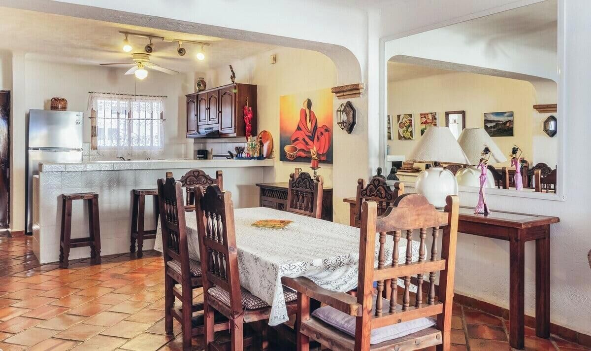 Un coin repas avec une table dressée pour quatre personnes, des chaises en bois, un ventilateur de plafond et une cuisine en arrière-plan.