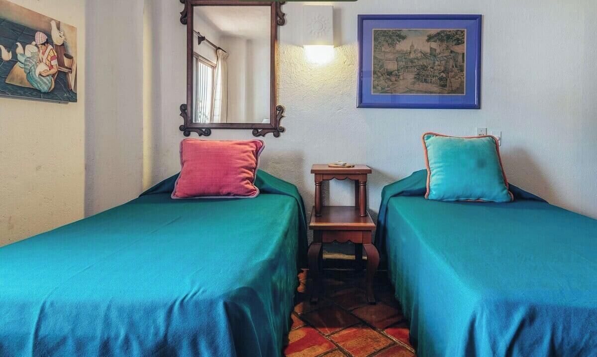 Una habitación con dos camas, una mesita, un espejo y obras de arte en las paredes.