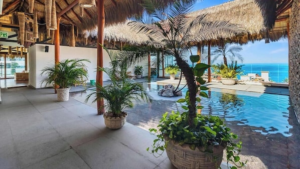An indoor pool area with a thatched roof, potted plants, and a view of the ocean. - Centric 1 BR Condo With Gym Amazing Rooftop Pool (Puerto Vallarta)