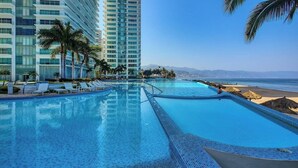 A swimming pool with lounge chairs, a beach, and a tall building in the background. - Luxury Oceanfront 2BR Condo With Private Balcony (Puerto Vallarta)