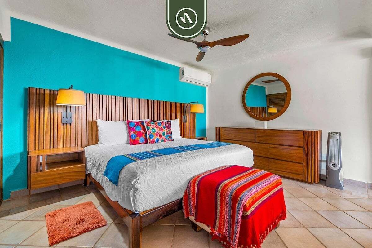 Une chambre d’hôtel avec une tête de lit en bois, un lit recouvert d’une courtepointe blanche et bleue, un plaid rouge, une table de chevet en bois avec une lampe, un miroir rond et un radiateur portatif.