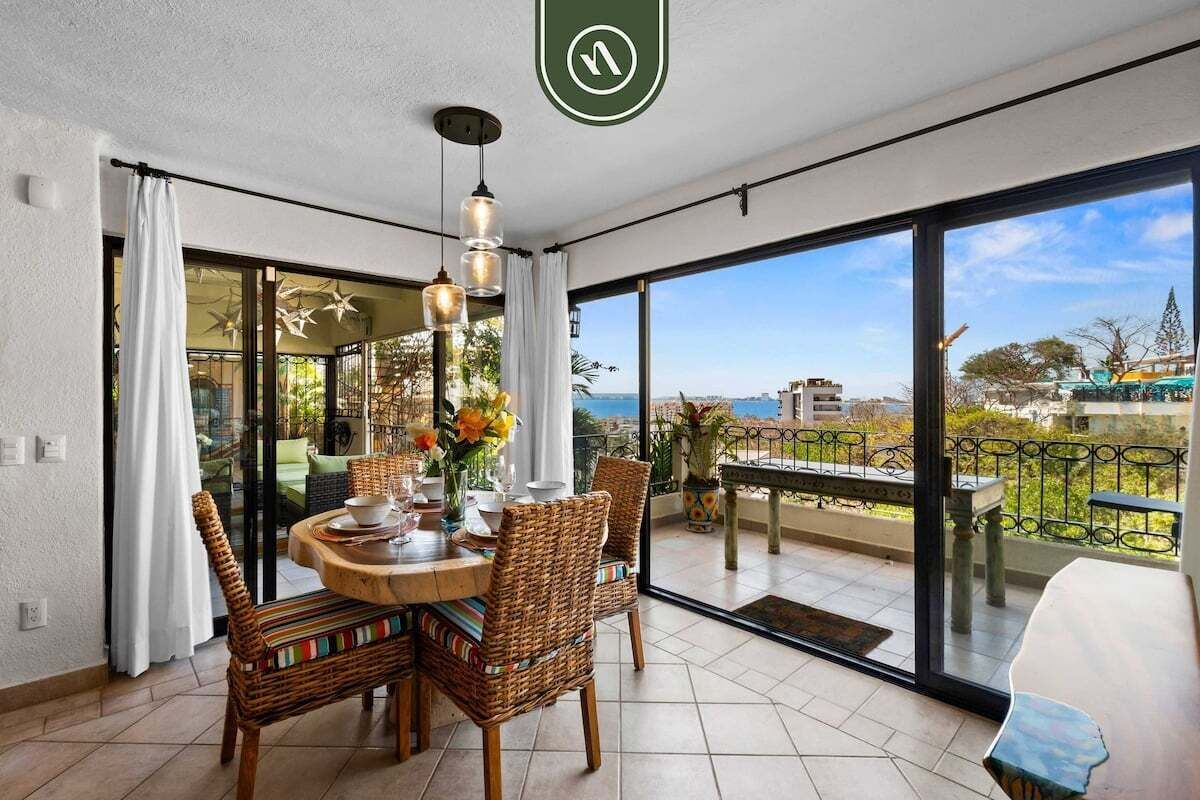 A dining area with wicker chairs and a round table, large sliding glass doors leading to a balcony with a view of the sea.