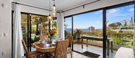 A dining area with wicker chairs and a round table, large sliding glass doors leading to a balcony with a view of the sea.