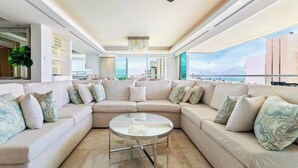 A modern living room with a large sectional sofa, a glass coffee table, and a view of the cityscape through large windows. - Luxury Oceanfront 4BR Condo - Pool - Ocean View (Puerto Vallarta)