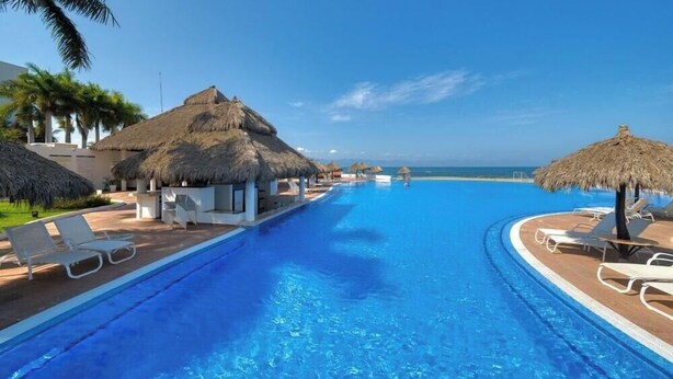 A clear blue pool with lounge chairs and thatched umbrellas, surrounded by palm trees and a clear sky. - Luxury Oceanfront 4BR Condo - Pool - Ocean View (Puerto Vallarta)
