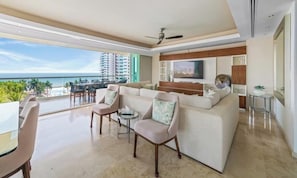 A modern living room with a large sofa, a coffee table, and a TV mounted on the wall. There is a balcony with a view of the ocean and palm trees. - Luxury Oceanfront 4BR Condo - Pool - Ocean View (Puerto Vallarta)