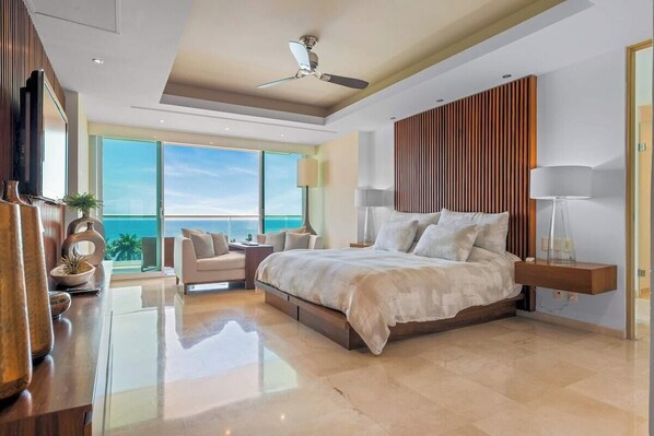 Un dormitorio moderno con una cama grande, ventilador de techo, televisor y vista al océano.