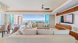 A modern living room with a large sofa, a TV, and a balcony with a view of the ocean. - Luxury Oceanfront 4BR Condo - Pool - Ocean View (Puerto Vallarta)