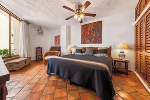 A bedroom with a bed, a ceiling fan, a nightstand, a lamp, a chair, a window with curtains, and a colorful painting on the wall. - Ocean View 2Br Condo-balcony-beach-pool - 802 (Puerto Vallarta)