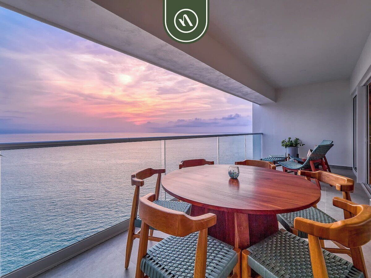 A round wooden table with chairs, a view of the sea, and a balcony with a railing.