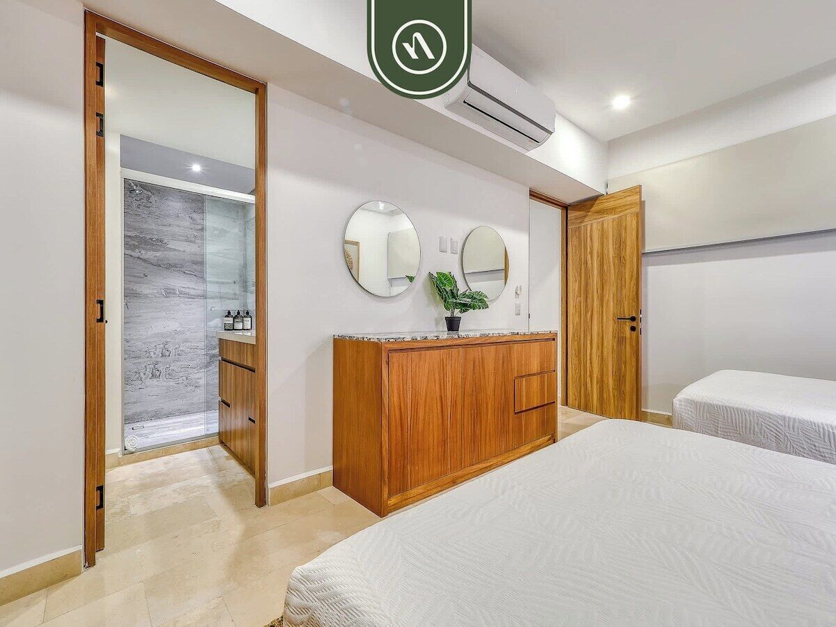 A bedroom with a wooden wardrobe, two bedside tables, a large bed, a mirror, and a door leading to a bathroom.
