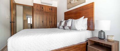 A bedroom with a wooden bed frame, white bedding, a bedside table with a lamp, and a framed picture on the wall.