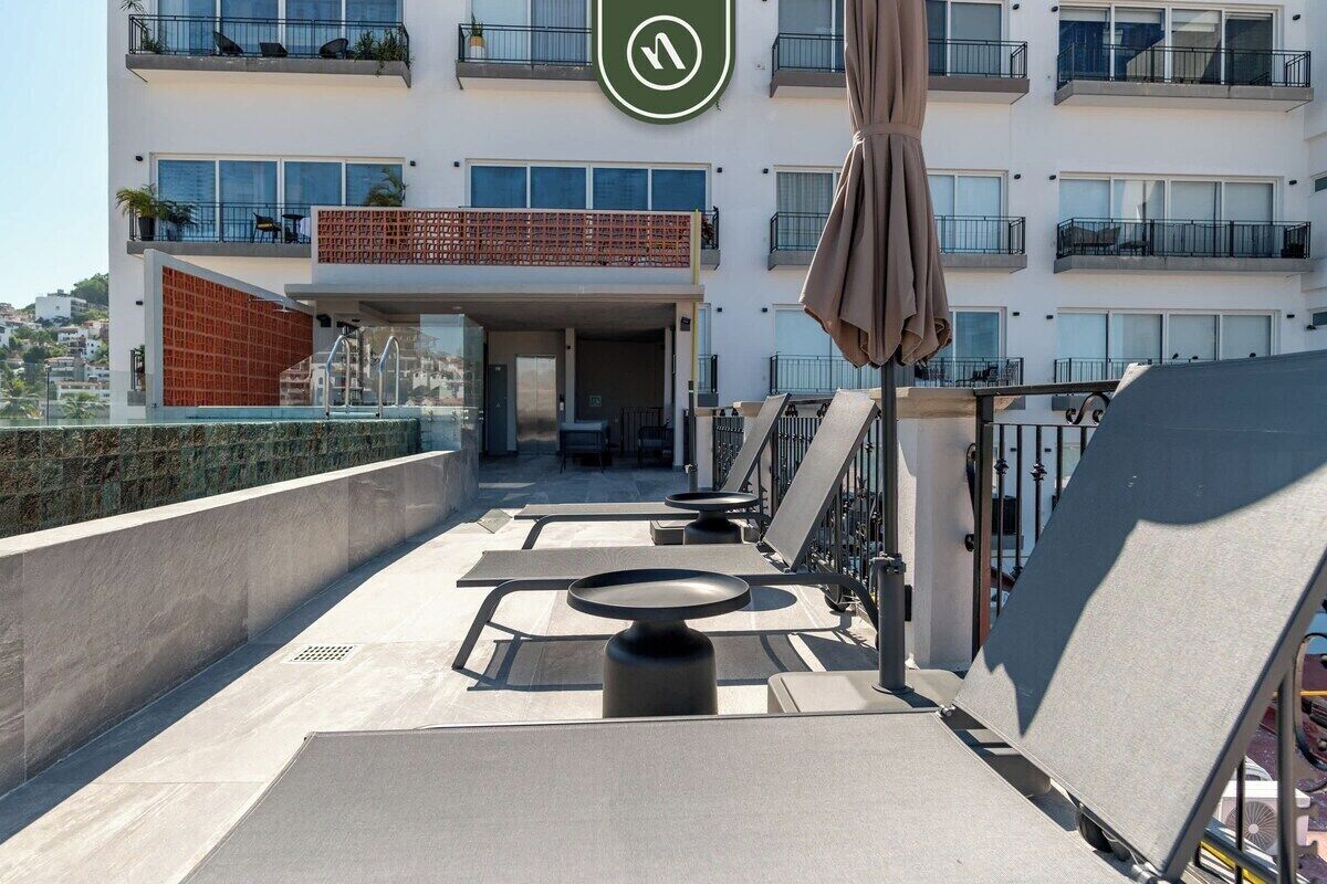 Una terraza en la azotea con mobiliario exterior, una zona cubierta y vistas a un edificio de varios pisos con balcones.
