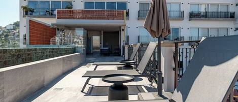 Una terraza en la azotea con mobiliario exterior, una zona cubierta y vistas a un edificio de varios pisos con balcones.