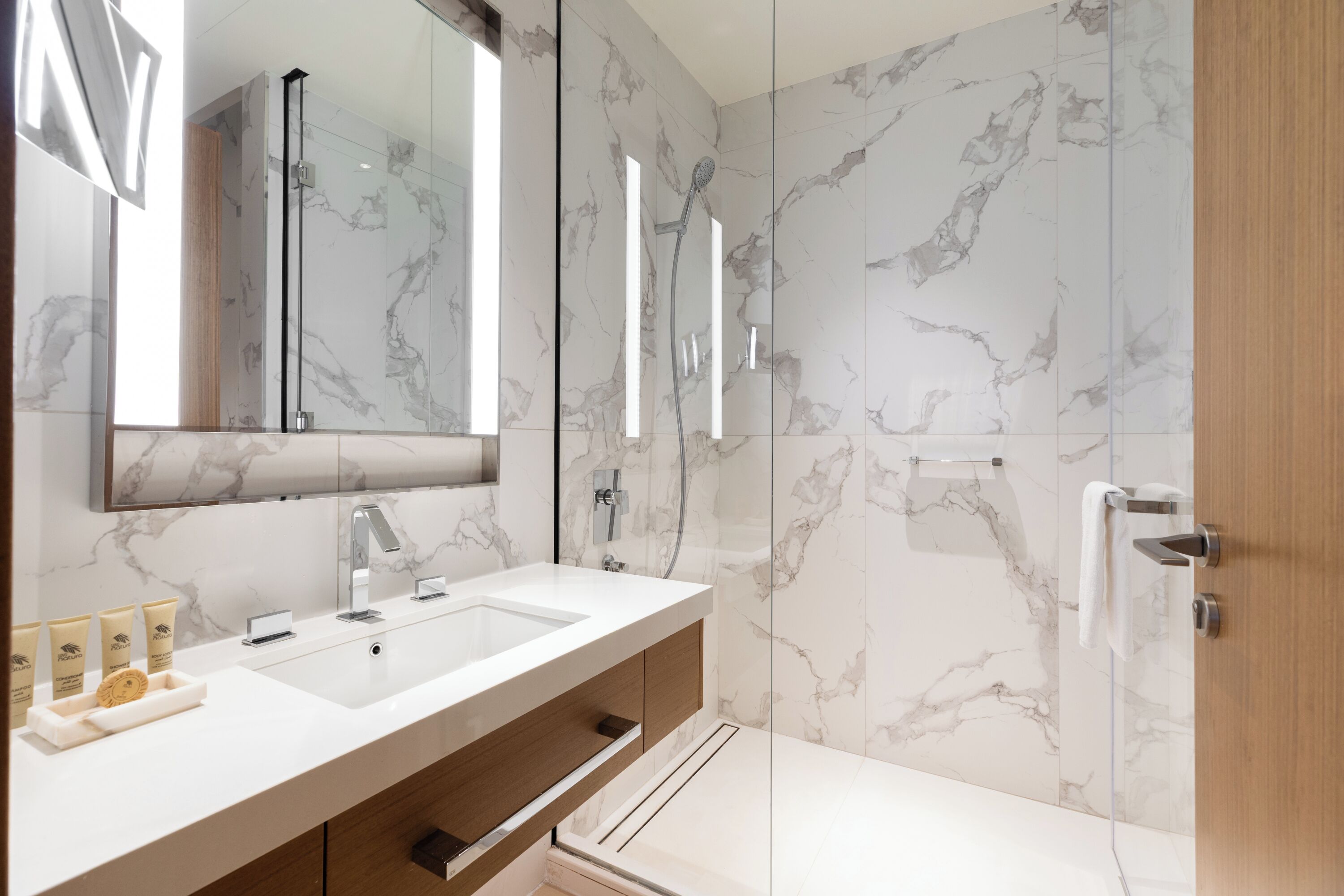A modern bathroom with a large mirror, two sinks, a shower, and marble walls.