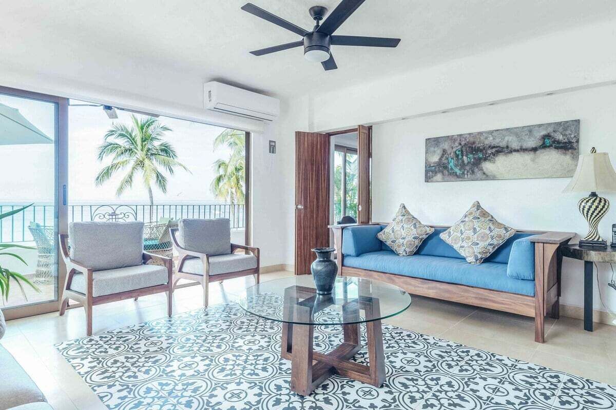 Een woonkamer met een glazen salontafel, een blauwe bank en stoelen, een plafondventilator en uitzicht op palmbomen.