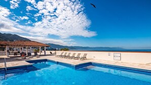 A swimming pool with lounge chairs and a covered area, overlooking a beach and mountains.