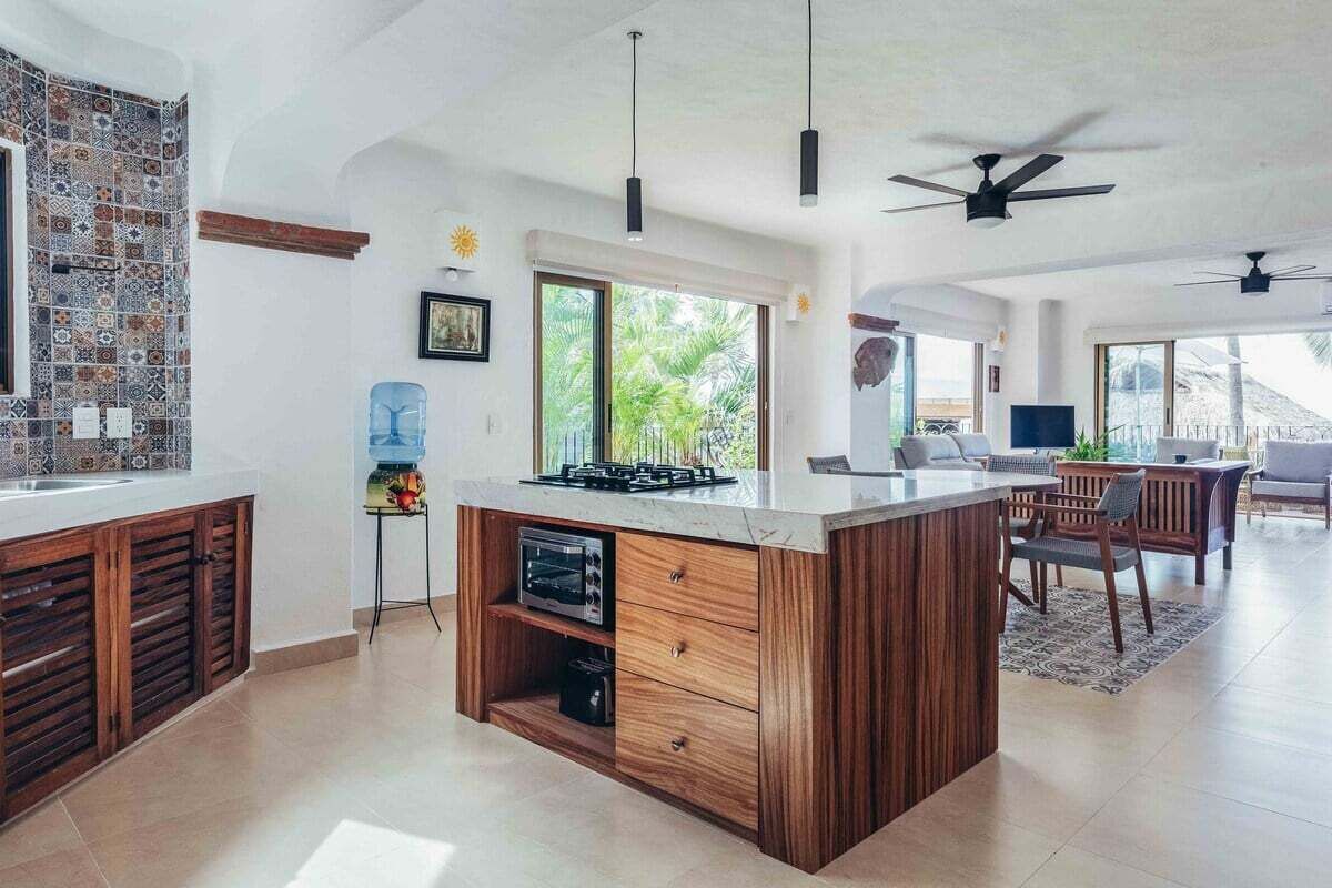Una cocina moderna con una isla central, armarios de madera y horno empotrado. Hay una zona de comedor con mesa y sillas, y un ventanal que ofrece vistas al exterior.