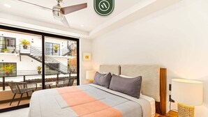 Une chambre d’hôtel moderne avec un grand lit, une table de chevet, une lampe et une vue sur le balcon extérieur, agrémenté de plantes et d’un escalier.