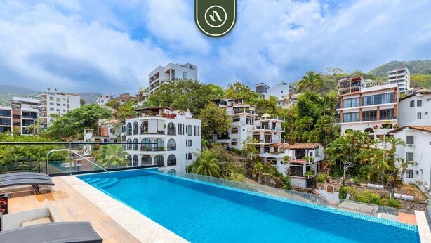 Une piscine entourée de chaises longues, elle-même bordée d’immeubles à plusieurs étages dotés de balcons et de verdure.