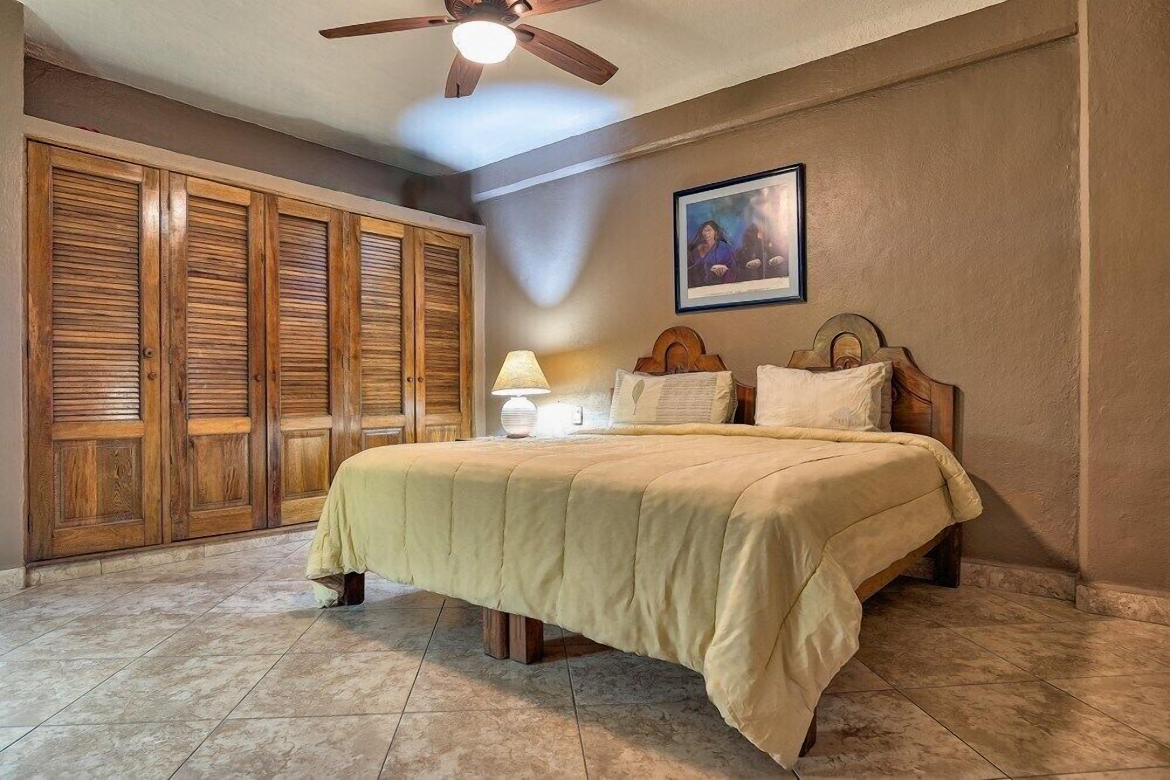 Une chambre à coucher avec un lit, un ventilateur de plafond, une table de chevet, une lampe et un tableau encadré au mur.