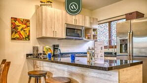 A modern kitchen with wooden cabinets, a stainless steel refrigerator, a microwave, and a coffee machine. There are two wooden stools and a colorful painting on the wall. - 1 BR Condo in Romantic Zone - Amazing Rooftop Pool (Puerto Vallarta)