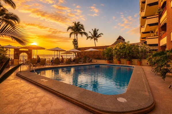 A swimming pool with lounge chairs and umbrellas, surrounded by palm trees and a building with balconies.