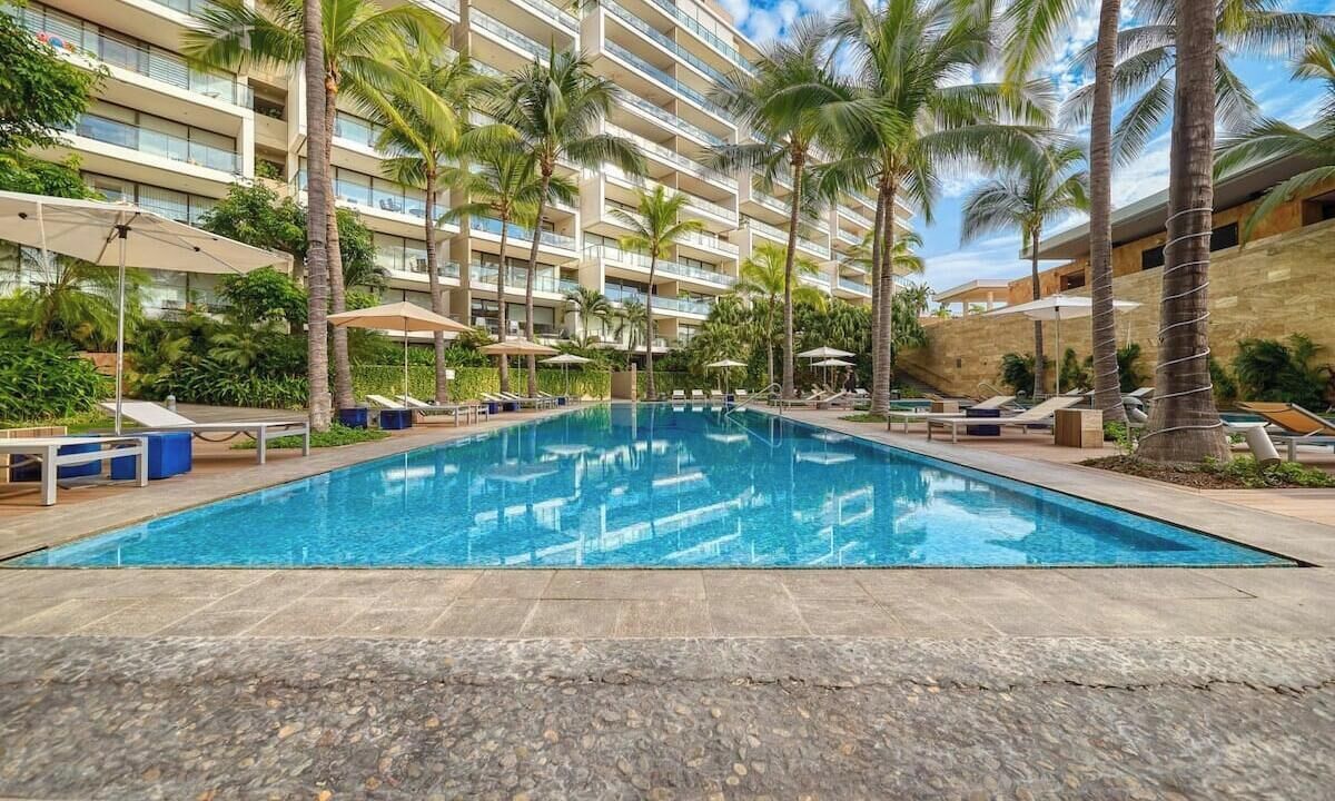 Ein Swimmingpool mit Liegestühlen und Sonnenschirmen, umgeben von Palmen und einem mehrstöckigen Gebäude.