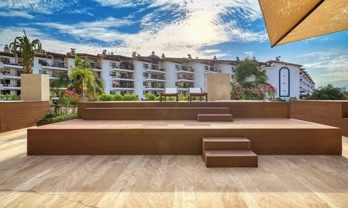 Un espacio exterior moderno con una terraza de madera, escalones que conducen a un área de descanso y vistas de un edificio de varios pisos con balcones.