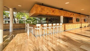 Un amplio bar con mostrador de madera y taburetes blancos, rodeado por un suelo pulido y grandes ventanales con vistas a un jardín tropical.