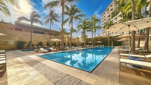 Una piscina rectangular con sillas de descanso y sombrillas, rodeada de palmeras y un muro de piedra.